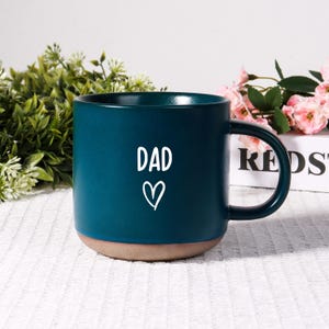 May include: A teal ceramic mug with the word "DAD" and a heart symbol in white. The mug has a brown base and a curved handle. The mug is on a white surface with greenery and pink flowers in the background.