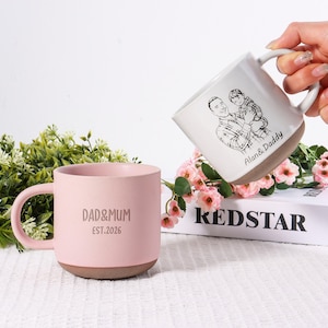 May include: Two ceramic mugs. One is light pink with "DAD&MUM EST.2026" printed on it. The other is white with a sketch of a man and child, and the text "Alan & Daddy". Both mugs have a brown base.
