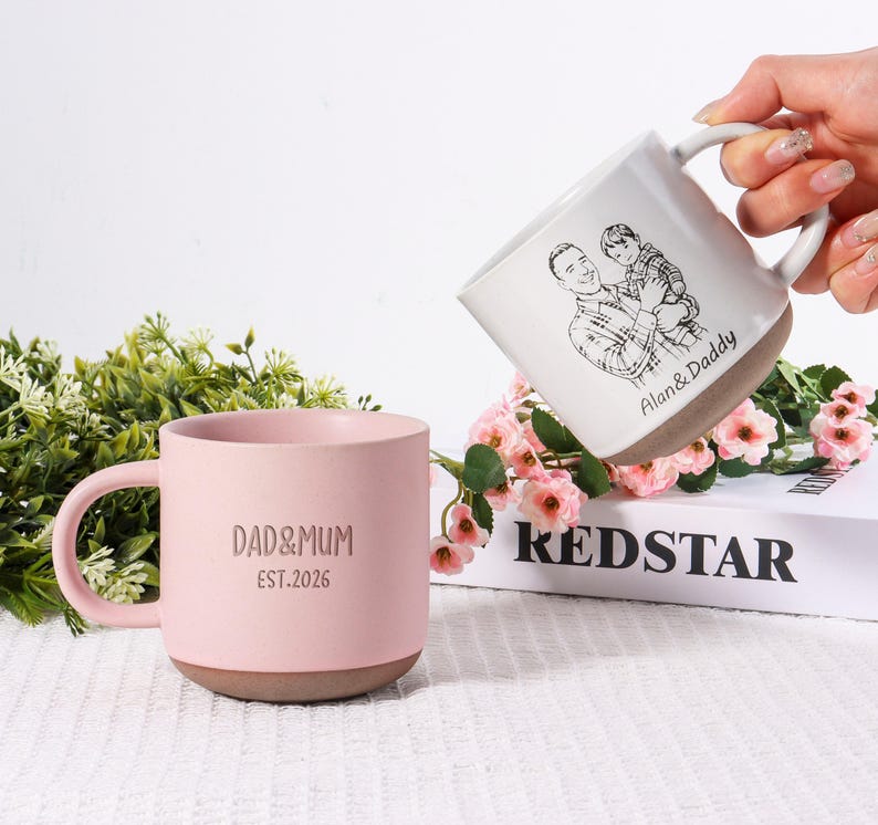 May include: Two ceramic mugs. One is light pink with "DAD&MUM EST.2026" printed on it. The other is white with a sketch of a man and child, and the text "Alan & Daddy". Both mugs have a brown base.