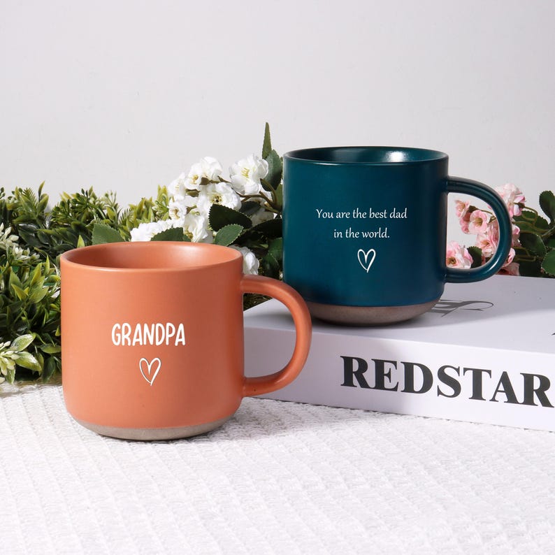 May include: Two ceramic mugs. One is orange with the word "GRANDPA" and a heart. The other is teal with the text "You are the best dad in the world." and a heart. Both mugs have a gray base.