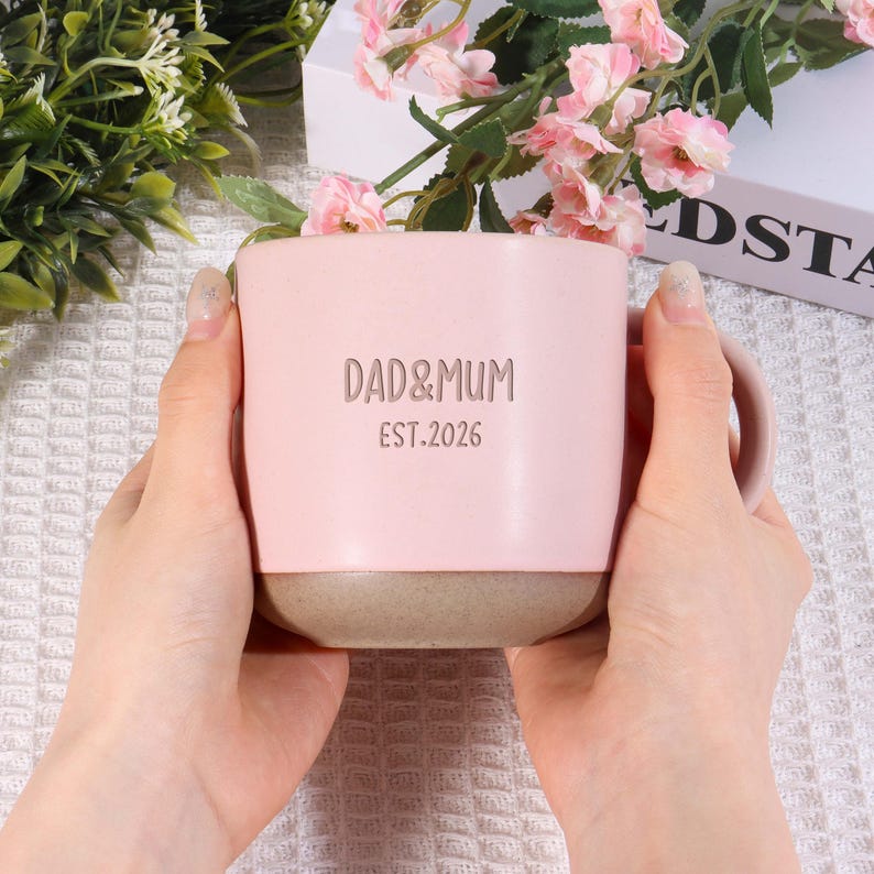 May include: A pink ceramic mug with the words "DAD&MUM EST.2026" printed on the front. The bottom of the mug is a neutral tan color. The mug is held by two hands.