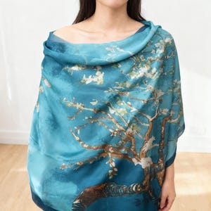 May include: A teal-colored scarf featuring a print of a tree with white blossoms, set against a blue background. The scarf is draped over the shoulders, showcasing the artwork's details and color palette. The design evokes a sense of art and nature.