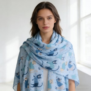 May include: A light blue scarf featuring a repeating pattern of stylized cats wearing sweaters. The cats are depicted in various poses, colored in shades of blue, white, and tan, interspersed with small white star-like shapes across the fabric.