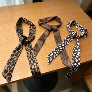 Puede incluir: Tres pañuelos de seda con estampados de animales. Uno es marrón con manchas negras, otro es marrón con manchas marrones oscuras y el tercero es blanco y negro. Los pañuelos están anudados.