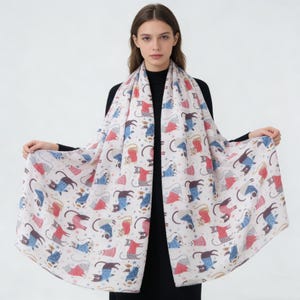 May include: A white scarf featuring a repeating pattern of stylized cats in various poses and outfits. The cats are depicted in shades of blue, red, and brown, with details like sweaters and hats. The scarf is draped, showcasing the full pattern.