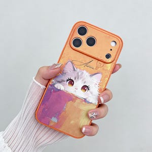 May include: Orange phone case featuring a cute cartoon cat peeking out of a colorful pocket. The case has a camera cutout and the word "Aurora" in script. The cat has big eyes and is white and gray.