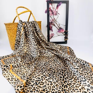 May include: A leopard print scarf with a gold border is draped over a woven basket. The scarf features a pattern of black spots on a tan background. A black framed art piece with pink flowers is in the background.