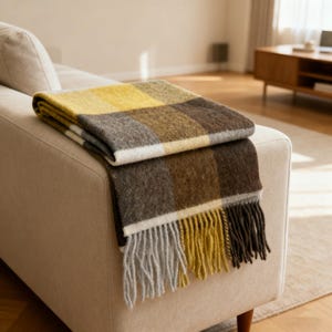 May include: A folded, checked throw blanket in shades of yellow, brown, and white rests on a beige sofa. The blanket has a fringed edge with yellow, grey, and brown tassels. The throw blanket is a cosy home decor item.