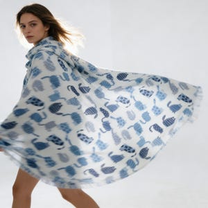 May include: A white and blue patterned scarf featuring a repeating cat design. The scarf is draped around a person's neck and shoulders, with the fabric flowing outward. The cat pattern includes various shades of blue.
