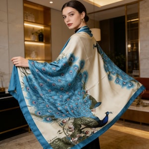 May include: A cream-coloured shawl with a peacock feather design in shades of blue and teal. The shawl has a teal border and features a full peacock illustration. The fabric appears to be soft and flowing.