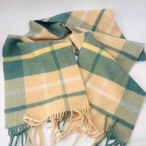 Puede incluir: Bufanda suave y tejida con un estampado de cuadros en tonos verde azulado, amarillo y crema. La bufanda tiene un borde con flecos y está hecha de un material cálido y acogedor. La bufanda está doblada y drapeada, mostrando el patrón.