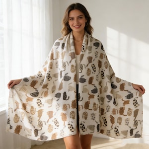 May include: A cream-colored shawl featuring a repeating pattern of stylized cats in shades of brown and black. The cats are depicted in various poses and patterns, creating a whimsical and cozy accessory. The shawl is draped over the shoulders.