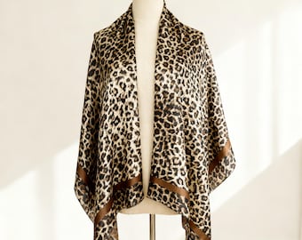 May include: A leopard print scarf draped over a mannequin. The scarf features a repeating pattern of black and brown spots on a tan background. A brown border frames the edges of the scarf, adding a touch of contrast.