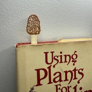 Puede incluir: Un marcapáginas artesanal con forma de seta marrón se inserta en un libro. El libro tiene una cubierta beige con el título "Using Plants For Healing" en letras rojas. El marcapáginas tiene forma de colmenilla.