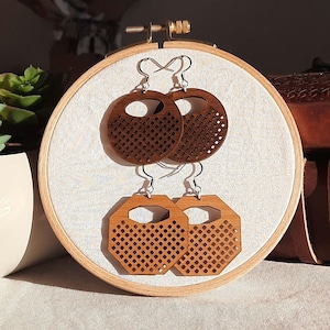 Könnte beinhalten: Ein Paar Holzohrringe mit geometrischem Design. Die Ohrringe sind braun und haben ein Kreuzstichmuster. Sie werden auf einem weißen Stickrahmen präsentiert.