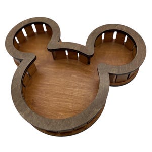 May include: Wooden Mickey Mouse-shaped tray with three compartments. The tray is made of light brown wood with a darker brown border. The tray's design features the iconic mouse ears and face shape.