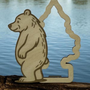 May include: A light brown wooden bear figure with a tree-shaped cutout. The bear has etched details and stands on a wooden base. The background shows a body of water.