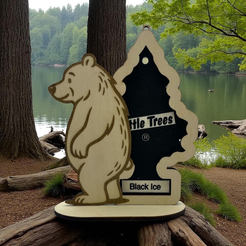 May include: Wooden bear and tree-shaped air freshener. The bear is light brown with a detailed outline. The tree-shaped freshener is black with the words "Little Trees" and "Black Ice" printed on it. The freshener is on a wooden base.