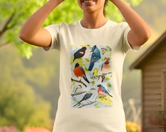 May include: Cream-colored t-shirt featuring a watercolor illustration of various birds perched on branches. The person in the image is holding binoculars, suggesting a birdwatching theme. The background is a blurred outdoor scene with greenery.