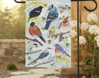 May include: A decorative garden flag featuring illustrations of various North American birds, including a Red-winged Blackbird, Blue Jay, and American Robin. The flag is displayed on a black metal garden stake, with a backdrop of natural elements.