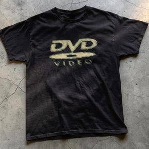 Op de afbeelding: Zwart T-shirt met een vervaagde DVD VIDEO-logo in een gepixelde, gebroken witte kleur. Het shirt met korte mouwen heeft een ronde hals en is gemaakt van zacht materiaal. Het shirt heeft een vintage stijl.