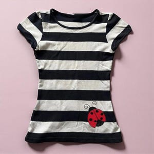Könnte beinhalten: Schwarz-weiß gestreiftes T-Shirt mit kurzen Ärmeln. Ein roter Marienkäfer mit schwarzen Punkten ist unten rechts aufgestickt. Das Shirt hat einen runden Ausschnitt und ist aus weichem Material.