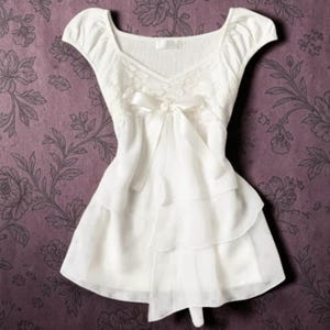 Op de afbeelding: Een witte blouse met korte mouwen, een ronde halslijn, een geribbeld lijfje en een transparante overlay. De voorzijde is in het midden versierd met een grote satijnen strik, die overgaat in een gelaagde, geplooide zoom voor een vloeiend silhouet.