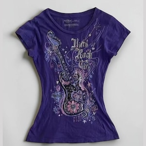 Könnte beinhalten: Ein lila T-Shirt mit einem dekorativen Gitarrendesign mit floralen und sternförmigen Akzenten. Das Shirt hat den Text "Hard Rock Girl" und "FIJI" aufgedruckt. Das Gitarrendesign ist in Rosa-, Lila- und Weißtönen gehalten.