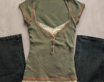 Puede incluir: Una camiseta de manga corta color verde oliva con escote redondo, ribete de encaje color crema, tapeta de botones y dobladillo superpuesto. La prenda se muestra junto a dos perneras de pantalón vaquero gris oscuro a cada lado.