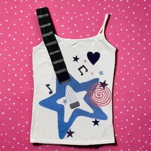 Op de afbeelding: Een wit hemdje met een grafisch ontwerp van een gitaarhals die uit een grote blauwe ster steekt. De print bevat een donkerblauw hart, muzieknoten, kleine sterren en een roze spiraalvormige cirkel. Het kledingstuk heeft dunne bandjes.