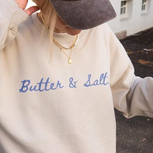 May include: A beige crewneck sweatshirt featuring the blue text Butter & Salt on the chest, paired with a dark brown baseball cap with white La Poésie Paris embroidery, a black shoulder bag, and a plastic cup containing an iced coffee beverage.