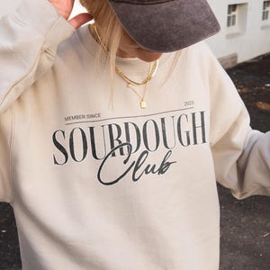 Może przedstawiać: Osoba nosząca brązową czapkę z daszkiem z napisem "La Paris" wyhaftowanym z przodu oraz kremową bluzę z nadrukiem "SOURDOUGH Club". Osoba trzyma czarną torebkę i napój kawowy ze słomką.