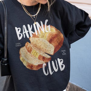 Può includere: Una felpa nera a girocollo con la grafica di una pagnotta affettata e il testo "BAKING CLUB", "EVERY SUNDAY" e "WAKE & BAKE". La felpa è indossata sopra una camicia bianca e una gonna di pizzo bianca, abbinata a un berretto da baseball grigio scuro.
