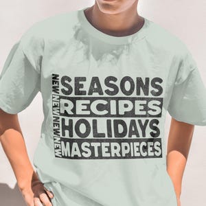 May include: A light green t-shirt with the words "SEASONS RECIPES HOLIDAYS MASTERPIECES" in black block letters. The word "NEW" is printed vertically on the left side of the shirt. The person is wearing sunglasses and brown pants.
