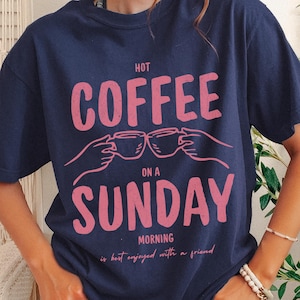 Può includere: Una maglietta blu navy con la scritta rosa "HOT COFFEE ON A SUNDAY MORNING is best enjoyed with a friend". La grafica include due mani che tengono tazze di caffè. La maglietta è etichettata "TRUE NAVY".