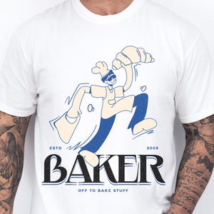 May include: White t-shirt featuring a cartoon baker with a croissant and a bag. The design includes the word "BAKER" in a bold font, with "ESTD 2006" above and "OFF TO BAKE STUFF" below. The shirt is a crew neck.