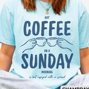 Può includere: T-shirt azzurra con la scritta "HOT COFFEE ON A SUNDAY MORNING is best enjoyed with a friend" in blu scuro. La maglietta presenta una grafica di due tazze di caffè. La parola "CHAMBRAY" è in basso.