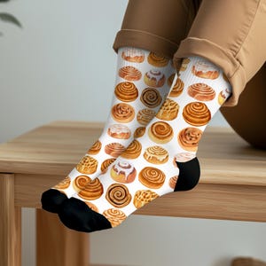 May include: White socks with a repeating pattern of watercolor cinnamon rolls. The socks have black heels and toes. The cinnamon rolls vary in size and frosting, with shades of brown and white.