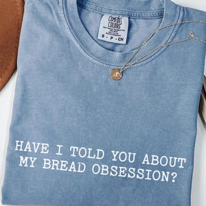 May include: A blue jean colored t-shirt with a crew neckline and a white graphic print that reads, HAVE I TOLD YOU ABOUT MY BREAD OBSESSION? The shirt features a Comfort Colors brand label at the inner neck and is paired with a gold tone pendant necklace.