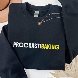 May include: Black sweatshirt with the text "PROCRASTIBAKING" in white and yellow. A gold necklace with a pendant is draped across the front. The word "BLACK" is visible in the bottom right corner.
