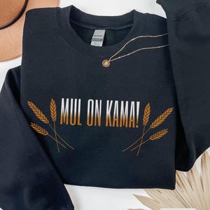 May include: Black crew neck sweatshirt with the text "MUL ON KAMA!" in white and gold, with wheat stalks on either side. A gold necklace with a pendant is draped across the sweatshirt. The word "BLACK" is in a white rectangle at the bottom.