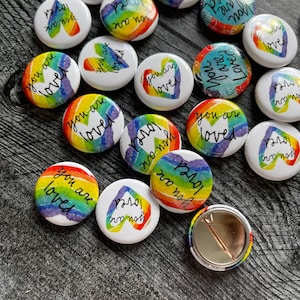 May include: A collection of rainbow-colored button pins with various messages, including "You are loved" and "Pride" with a rainbow heart.