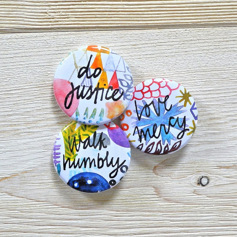 May include: Three colorful round buttons with inspirational messages. The first button says "do justice" in black script on a white background with watercolor-like designs in shades of pink, blue, and green. The second button says "love mercy" in black script on a white background with watercolor-like designs in shades of red, blue, and green. The third button says "walk humbly" in black script on a white background with watercolor-like designs in shades of purple, blue, and green.