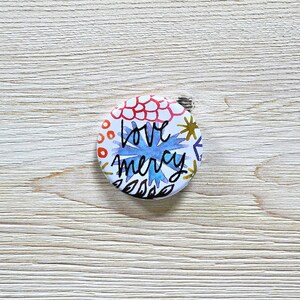 May include: A white button with a colorful floral design and the text "love mercy" in black.