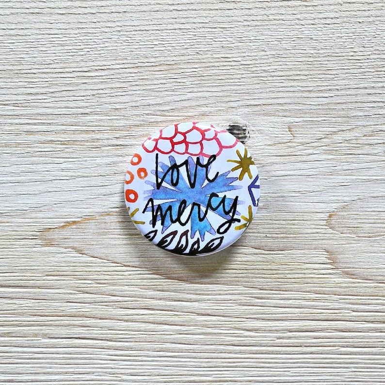 May include: A white button with a colorful floral design and the text "love mercy" in black.