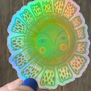 May include: A holographic sticker featuring a stylized sun with a smiling face. The sun has a green and yellow gradient with a scalloped edge. The sun's rays are detailed with dotted patterns.