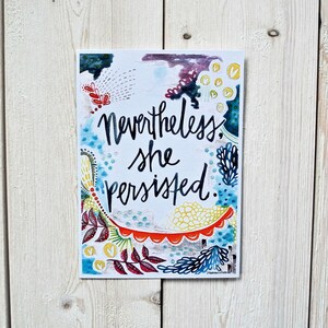 以下が含まれることがあります： カラフルな抽象的な背景と「Nevertheless, she persisted.」というテキストが書かれた水彩画のプリントです。