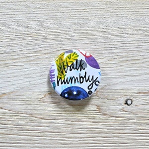 May include: A round button with a white background and colorful abstract designs. The text "Walk humbly" is written in black on the button.