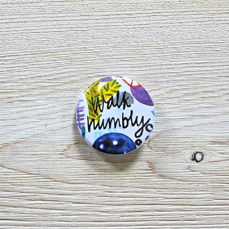 May include: A round button with a white background and colorful abstract designs. The text "Walk humbly" is written in black on the button.