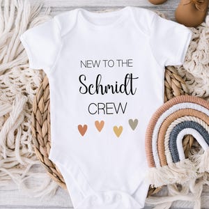 May include: White baby onesie with the text "New to the Schmidt Crew" in black. Five small hearts in different shades of brown, orange, and gray are below the text.